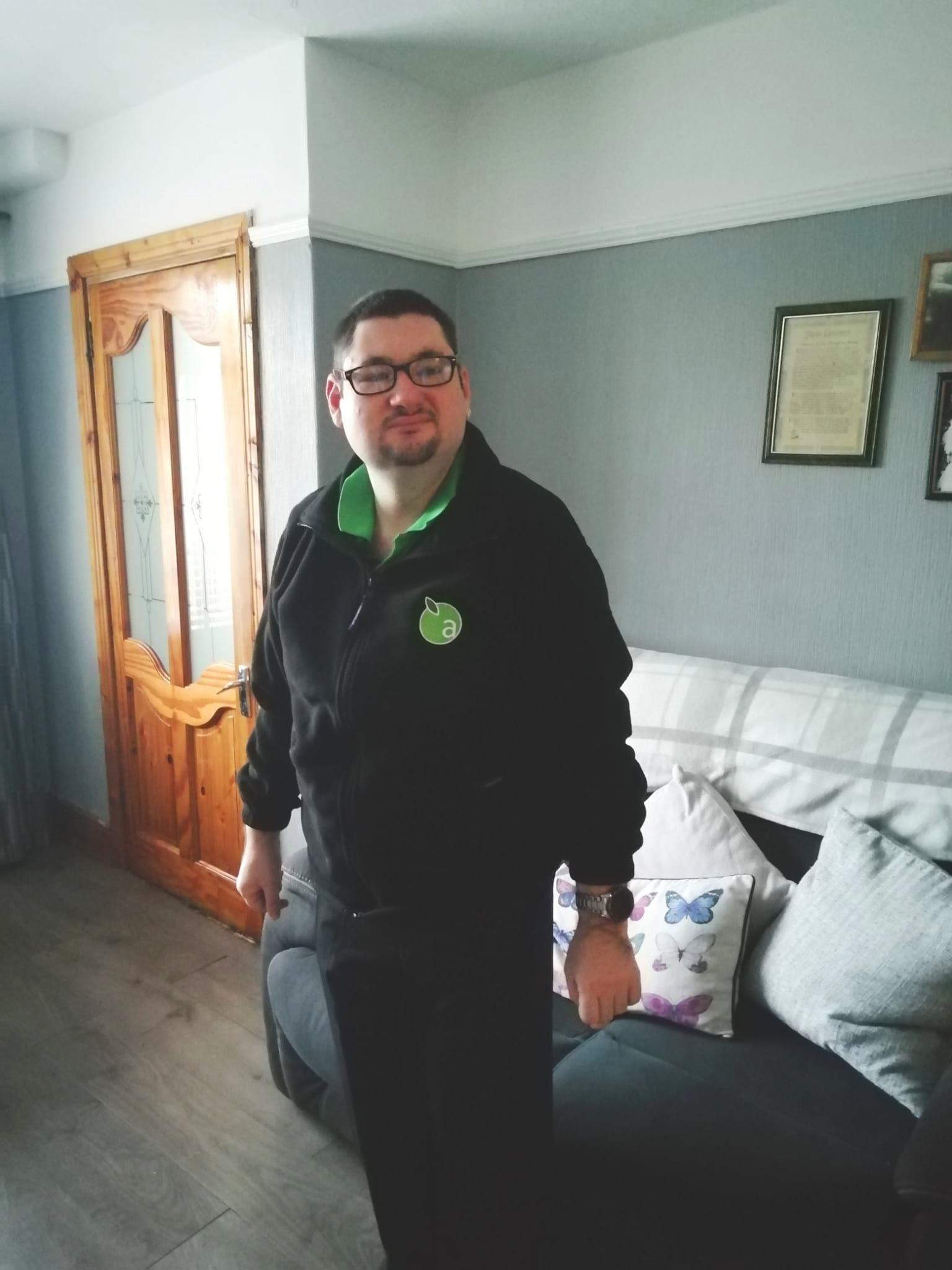 The image shows Luke Mannering getting ready for a shift at Applegree, wearing his black Applegreen uniform, he is standing in his living room, smiling. 