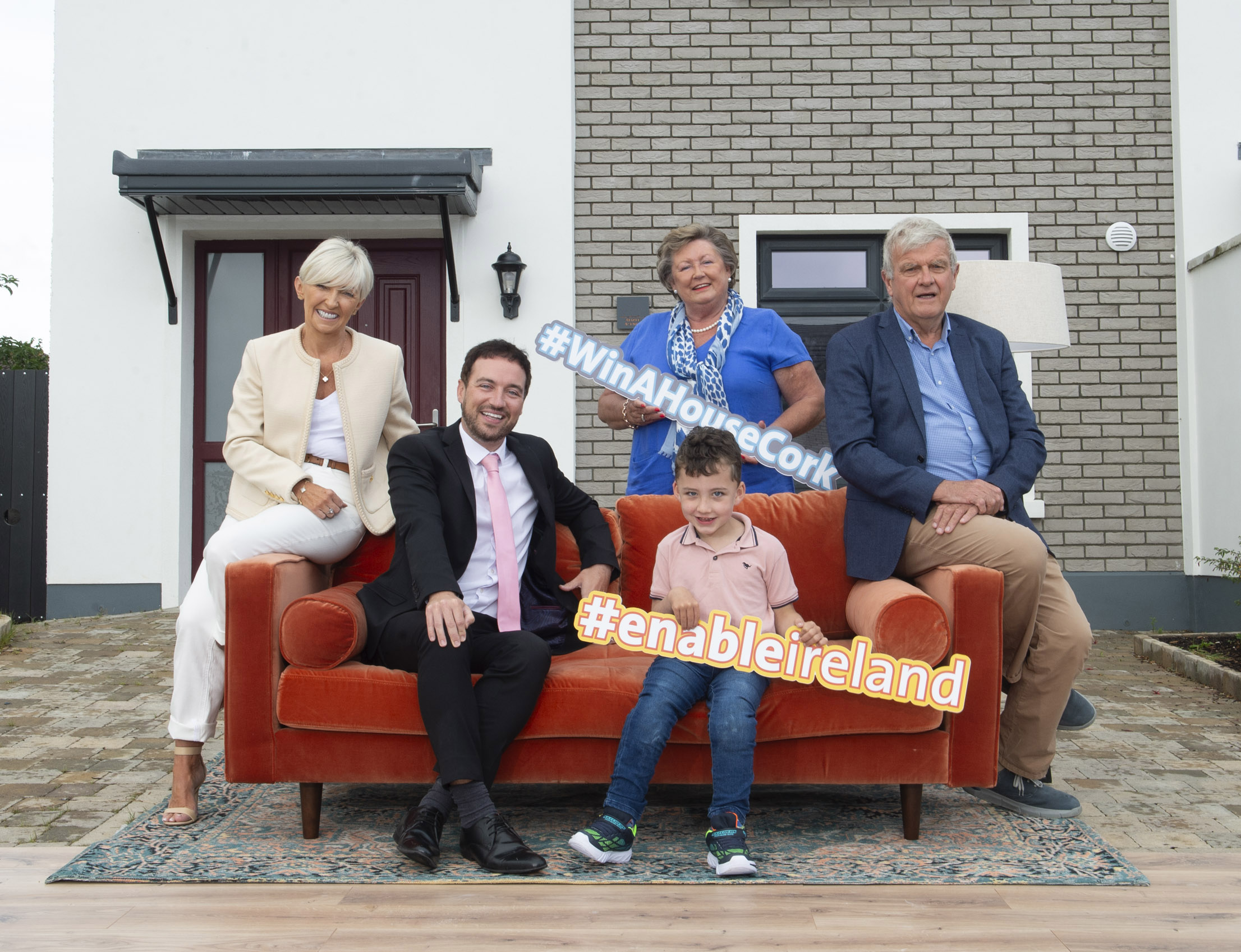 Four adults and a child sit on a couch in front of a house. They hold signs saying #winahouse and #enableireland