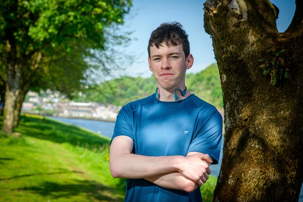 A man in a blue t-shirt standing next to a tree with his arms crossed.