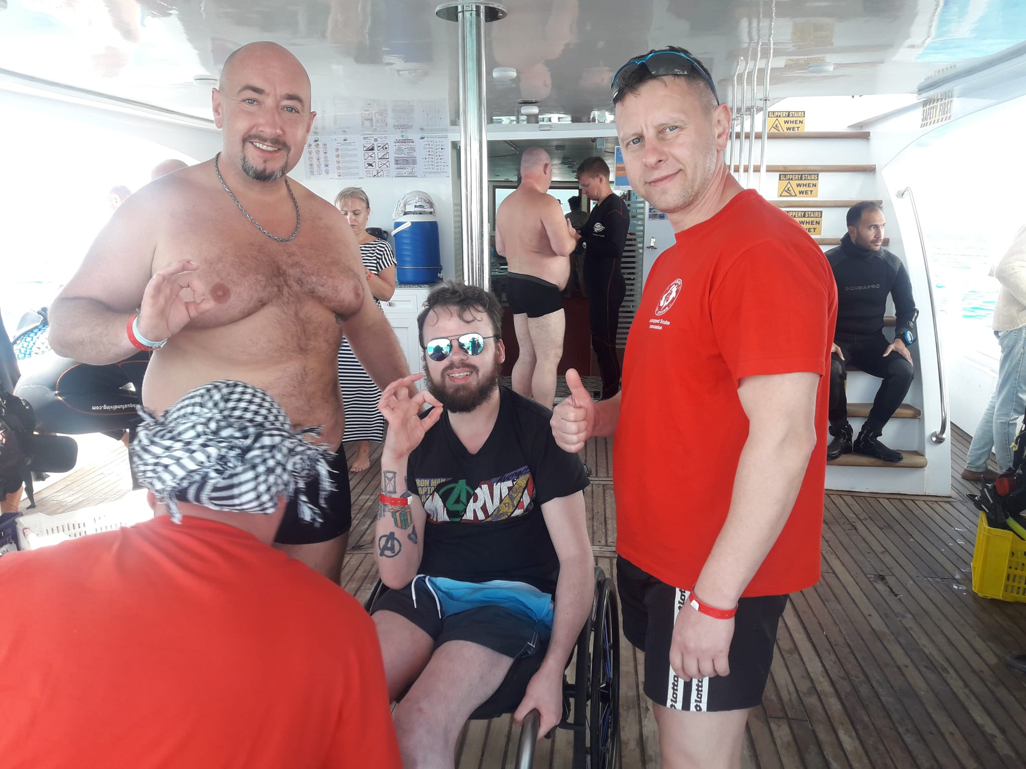 A young man using a wheelchair on a boat with two men standing next to him. They are all doing the 'okay' hand signal.