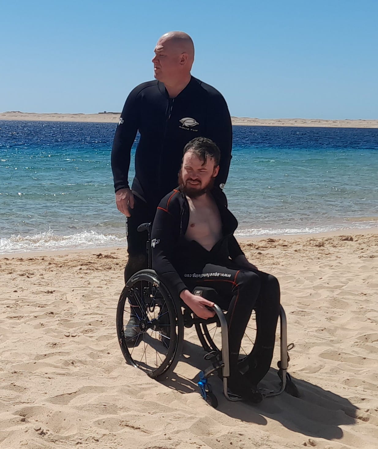 A photo of a young man dressed in a wet suit using a wheelchair on the beach with the sea behind him.