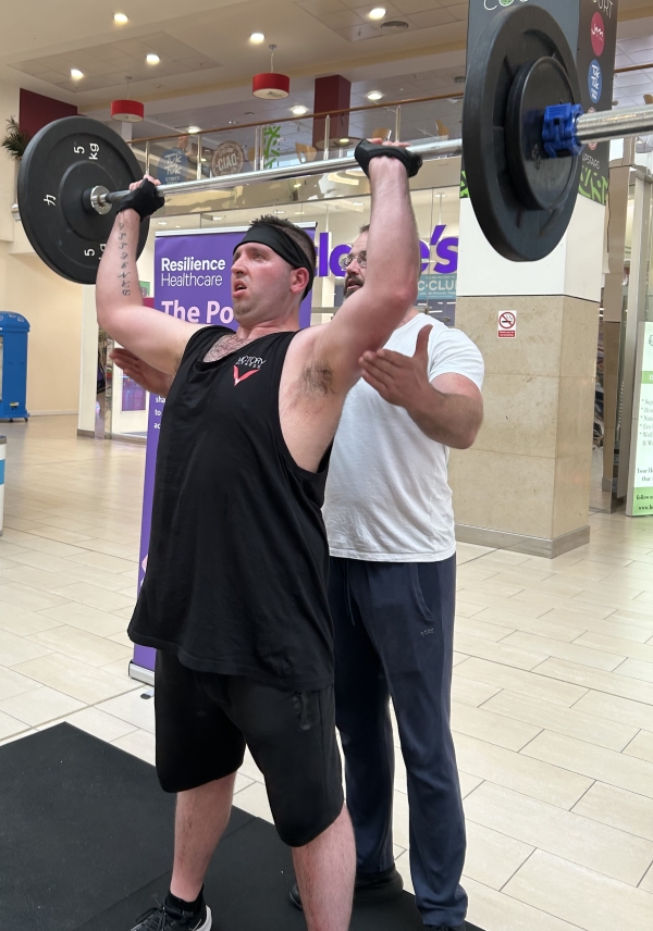 A photo of a man lifting a weight bar over his head. Another man stands behind him for support.