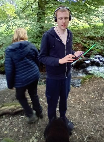 A photo of a young man standing in a forest, wearing headphones and holding an electronic device.