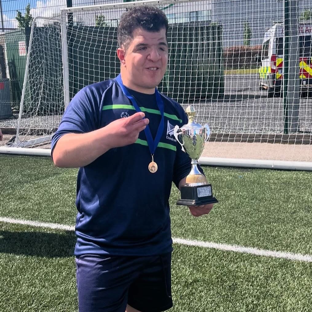 A person wearing a navy football uniform stood next to a football goal net wearing a medal and holding a trophy