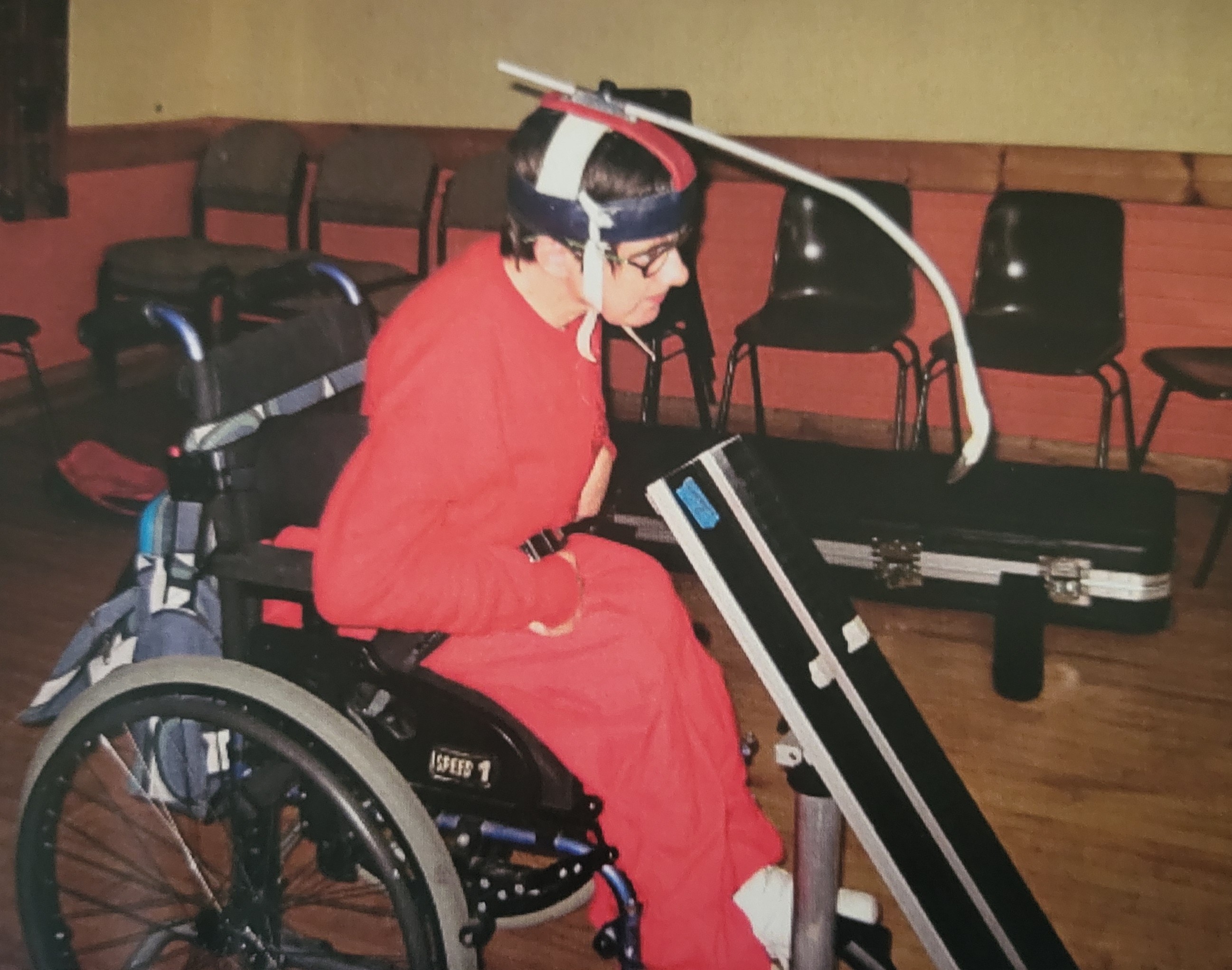 Person in a wheelchair wearing a red outfit and a head-mounted device with an attached white tube in a room with chairs and a wooden floor.