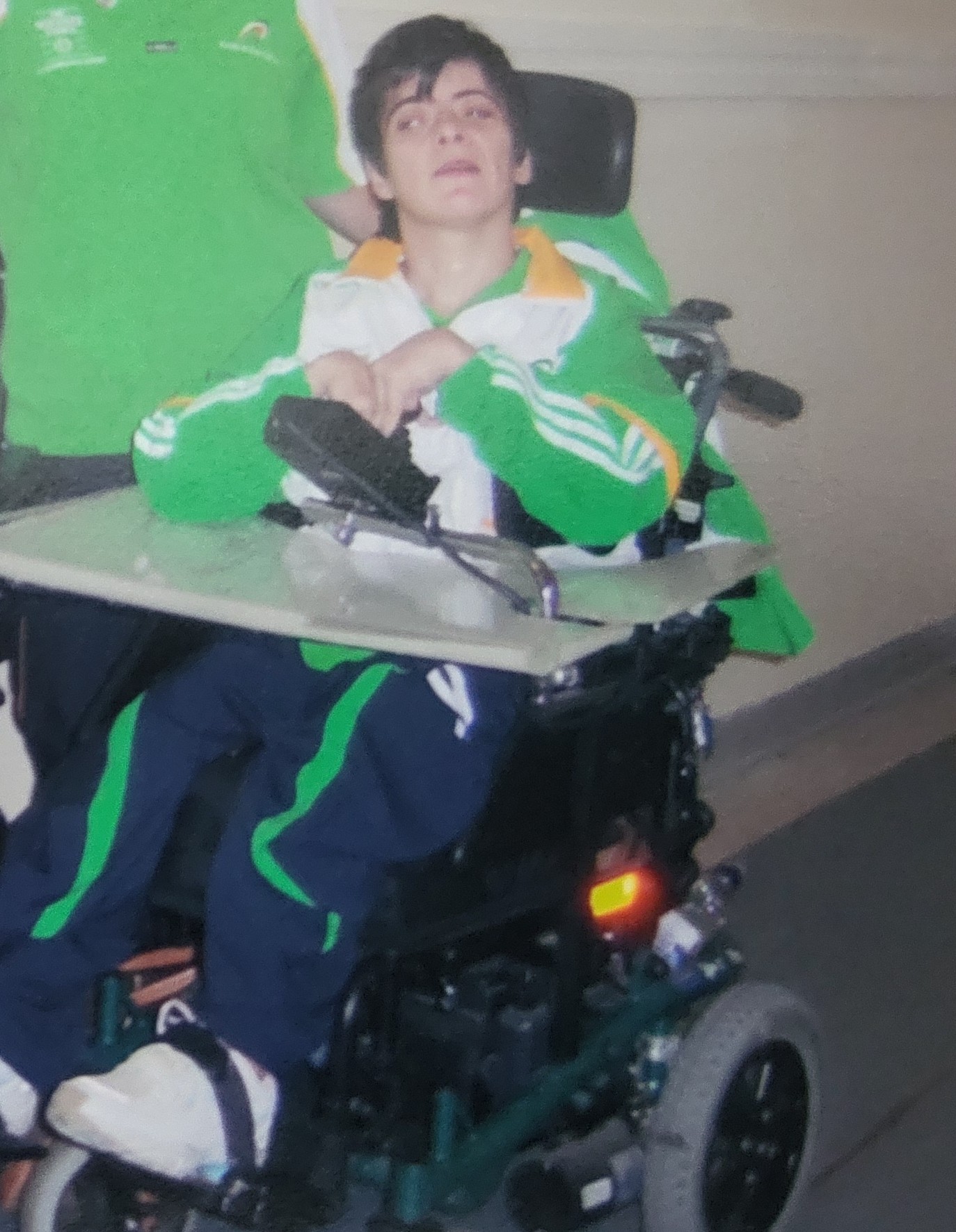 Person wearing a green and white jacket with yellow accents seated in a motorized wheelchair with a tray table attached.