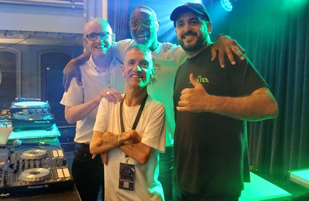 Four men posing together near DJ equipment in a club with green stage lights and a blue spotlight behind them.