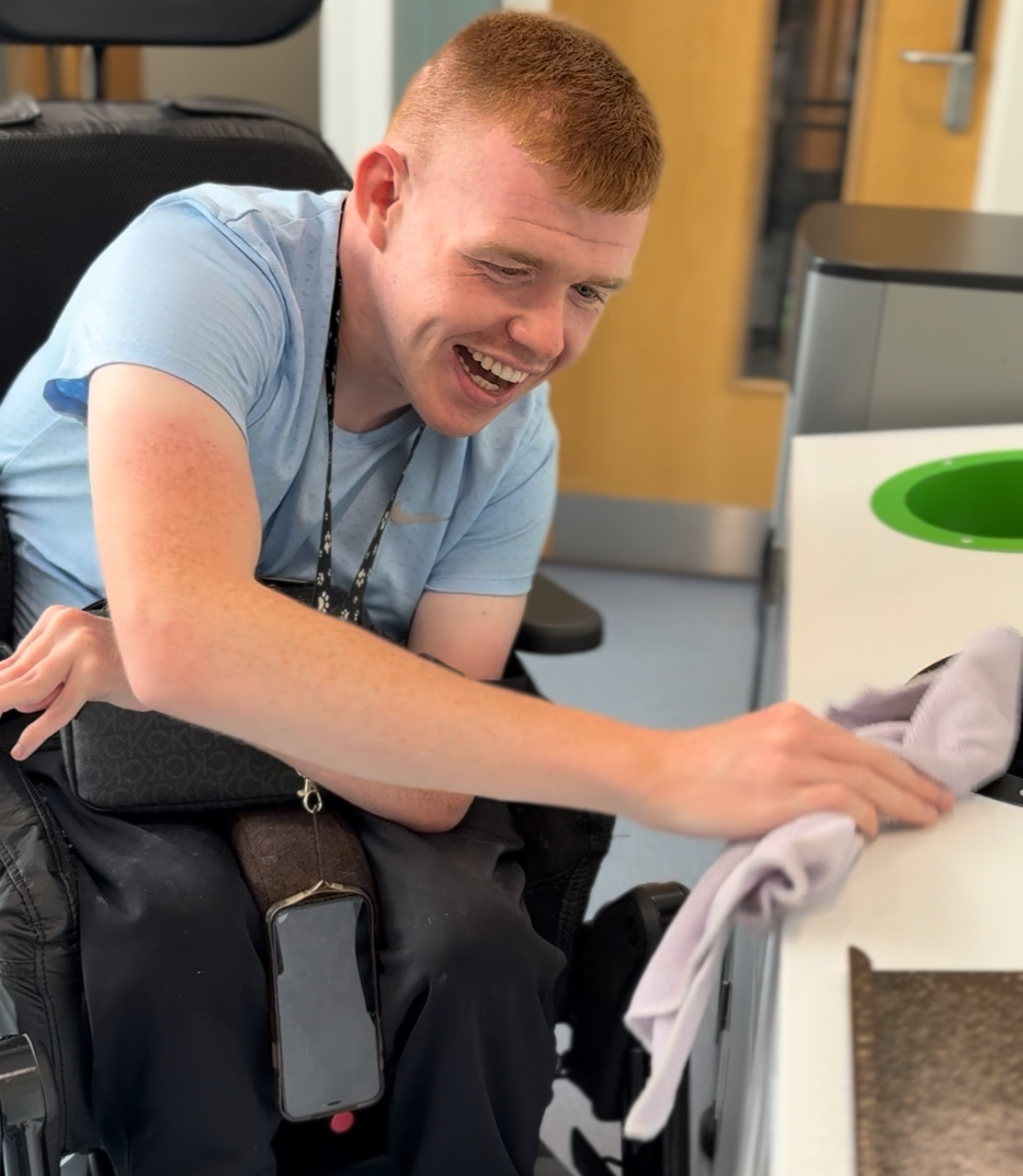 Person in a wheelchair wearing a light blue shirt cleaning a white countertop with a cloth in an indoor setting.