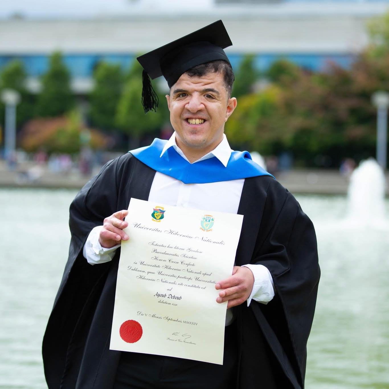 A person, outside by a fountain, wearing a graduation gown and mortarboard, holding a certificate.