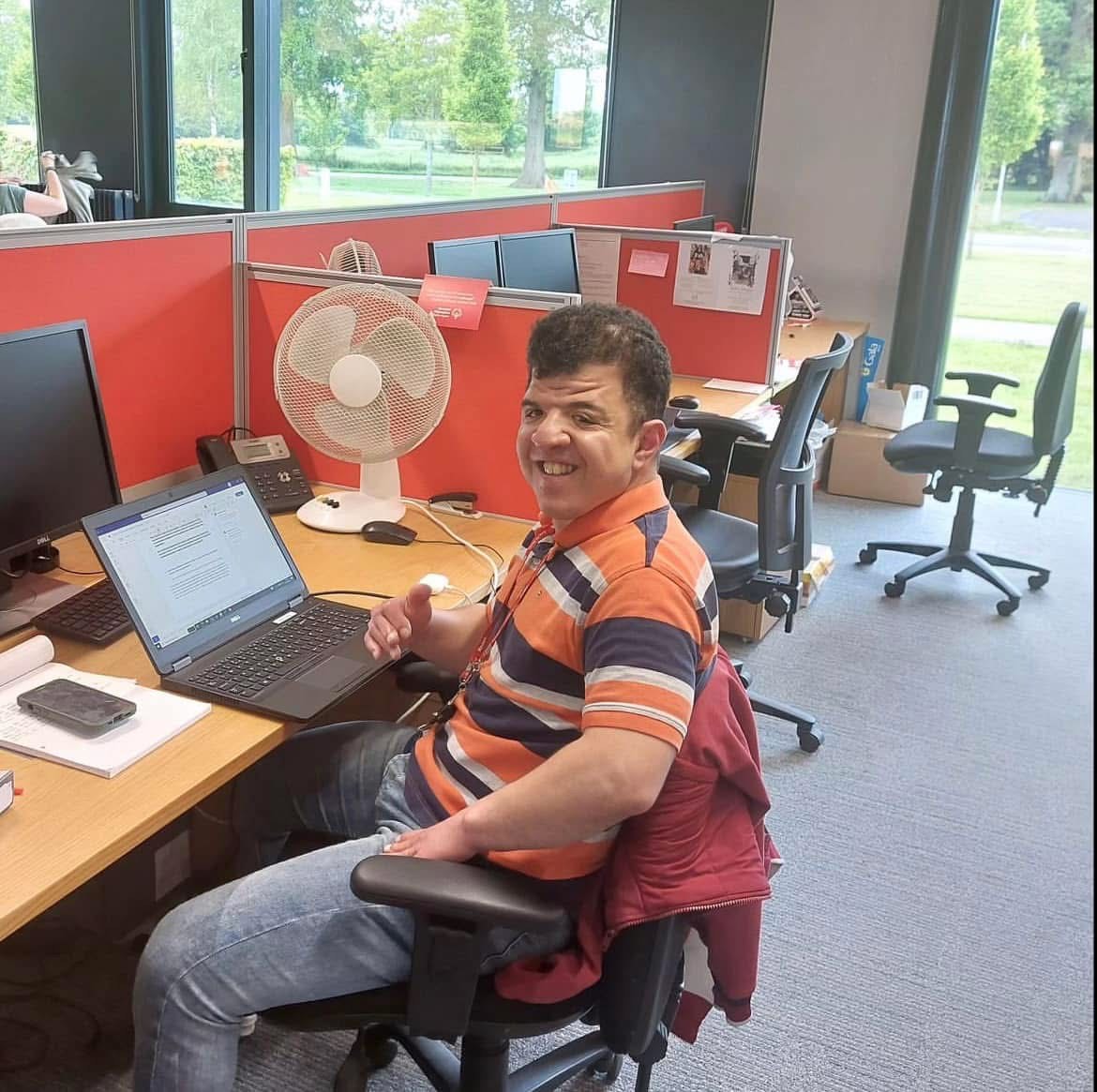 Man in a striped orange and navy shirt sitting at a desk with a laptop in a bright office cubicle with red dividers and a white fan.