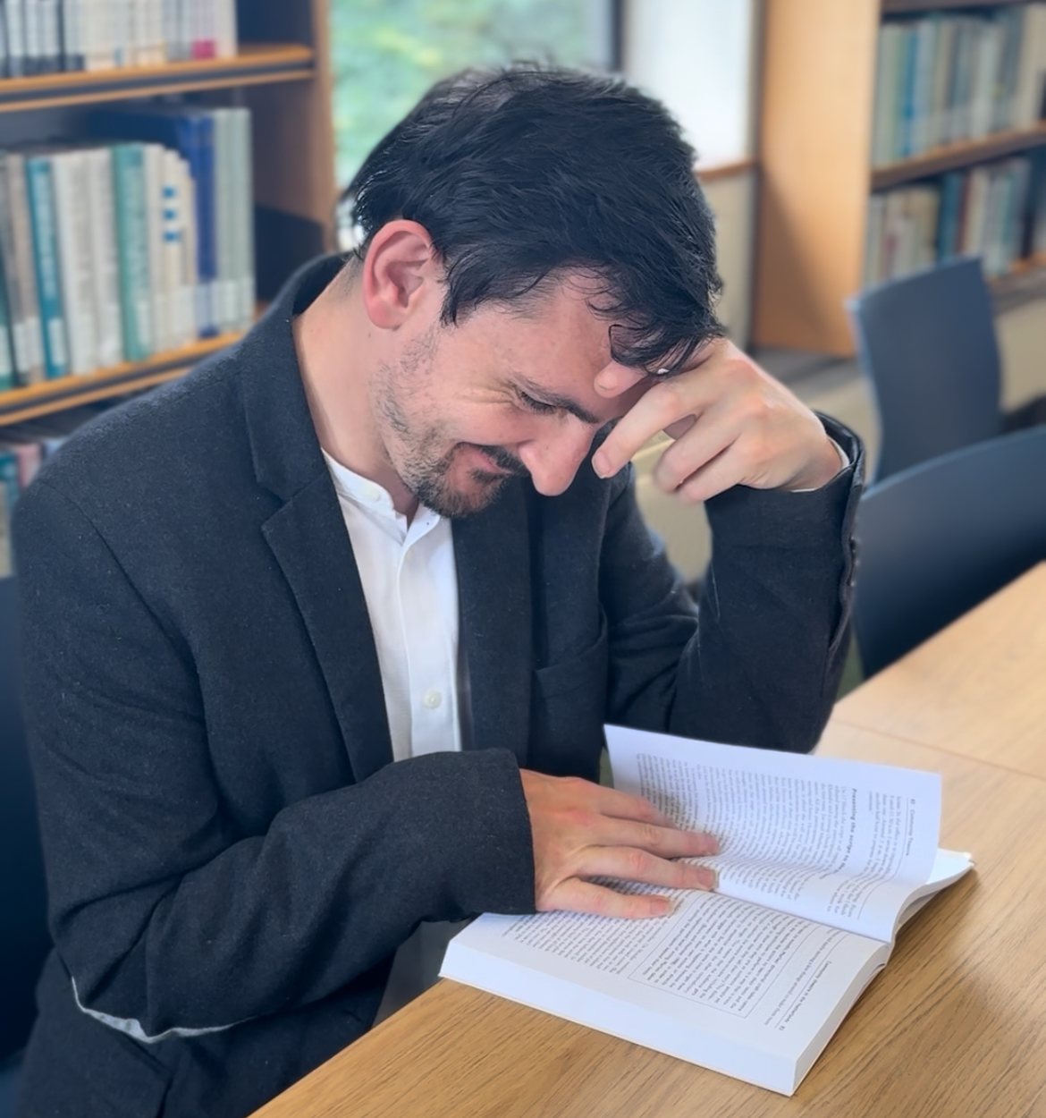 A young man in a library sat a table with his head resting on his hand. He is reading a book which is laying on the table.