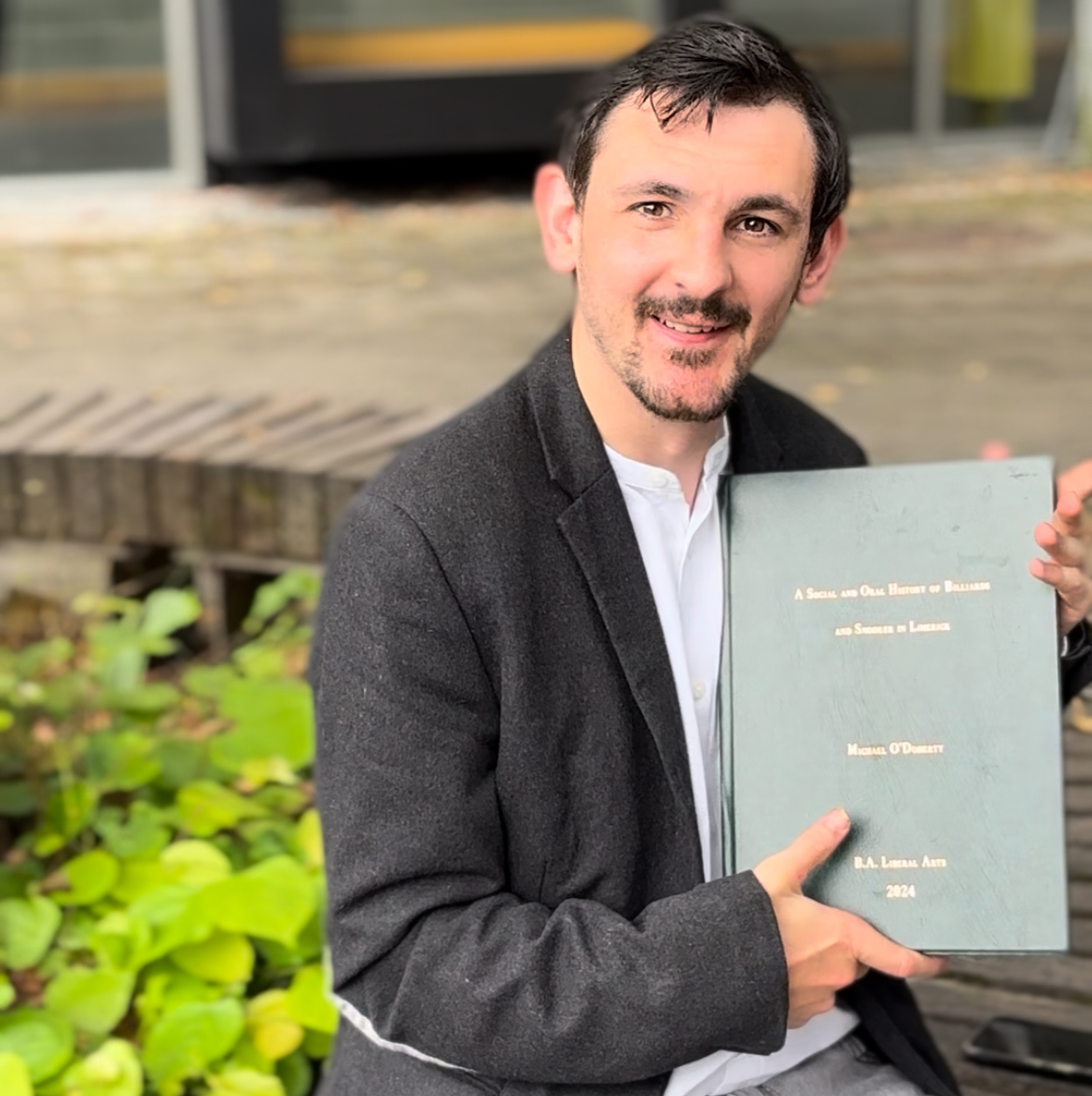 Person, sitting outdoors, in a dark blazer holding a green hardcover thesis titled 'A Social and Oral History of Billiards and Snooker in Limerick' .