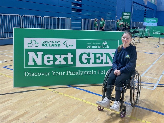 Photo of a young girl wearing a navy hoody and using a wheelchair next to large green Paralympics Ireland 'Next Gen' sign.