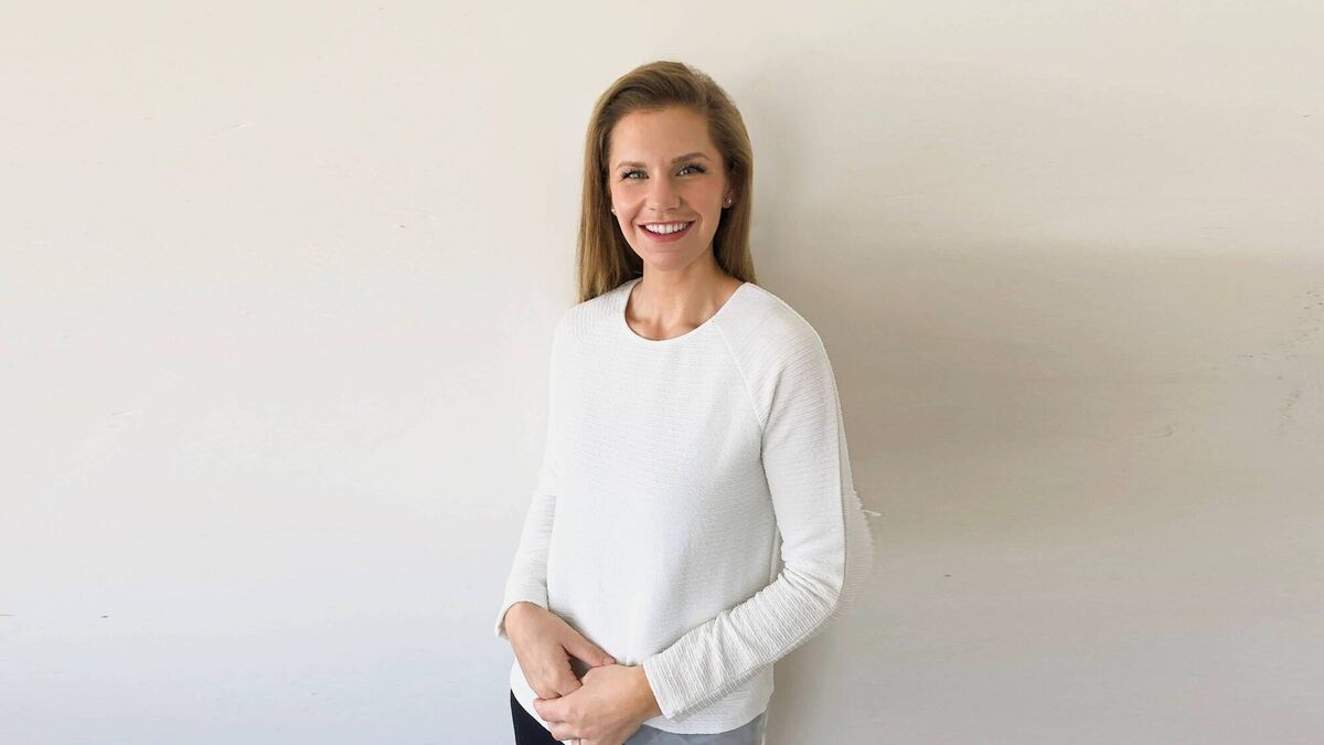 A person standing against a plain light‑coloured wall, wearing a long‑sleeved white top and dark trousers with hands loosely clasped in front.