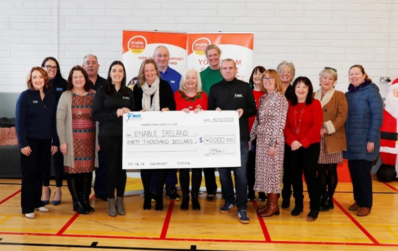 Group of people standing in a sports hall holding a large donation cheque made out to Enable Ireland for $40,000, with Enable Ireland banners displayed in the background.