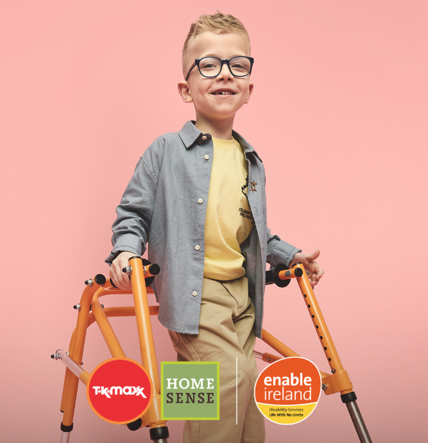 Child using an orange walker with large wheels against a solid pink background, dressed in beige pants and a blue shirt.