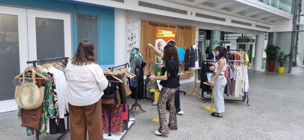 People browsing clothing racks at a pop‑up fashion sale in a bright indoor venue, with outfits, accessories, and display stands set up along a walkway under a “Happy Social Week” notice board.