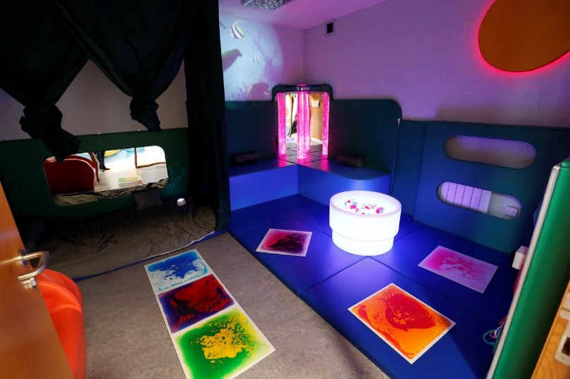 Multi‑sensory therapy room with soft padded seating, colourful illuminated floor panels, a light‑up bubble tube, and interactive wall features designed for sensory stimulation.