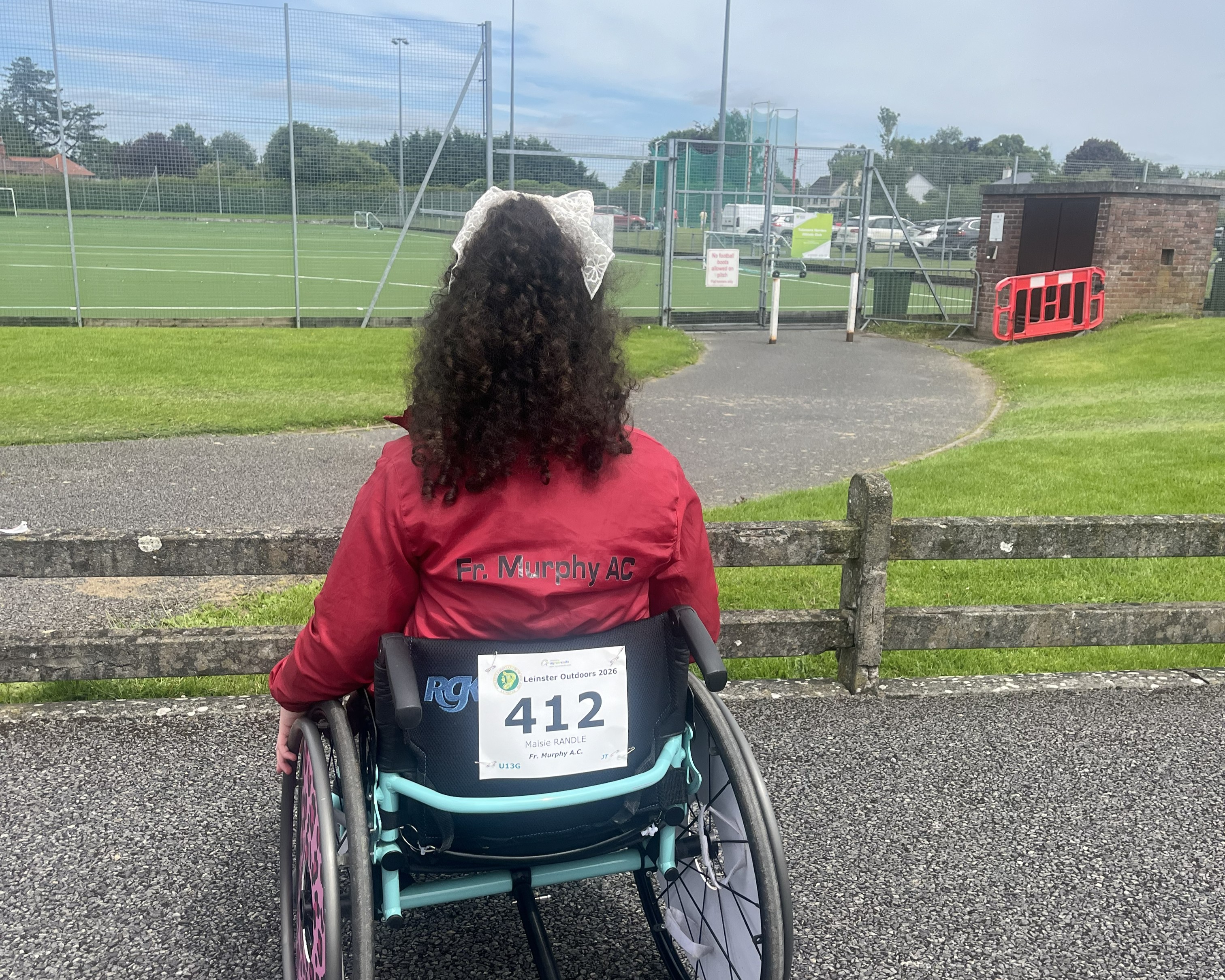 Life With No Limits Champion Maisie Randle, an international gold medallist, seated in a wheelchair beside an athletics track, looking towards the competition area while wearing a Fr. Murphy AC jacket and race number at a Leinster Outdoor athletics event.