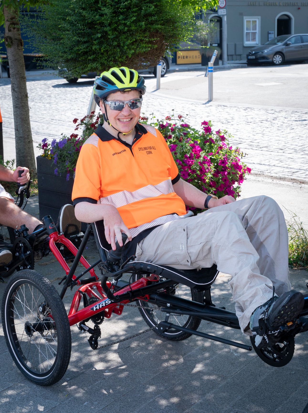 Life With No Limits Champion Kevin riding an adapted recumbent bicycle during the Cycle for All Cork programme, wearing a helmet and high‑visibility clothing beside colourful flowers.
