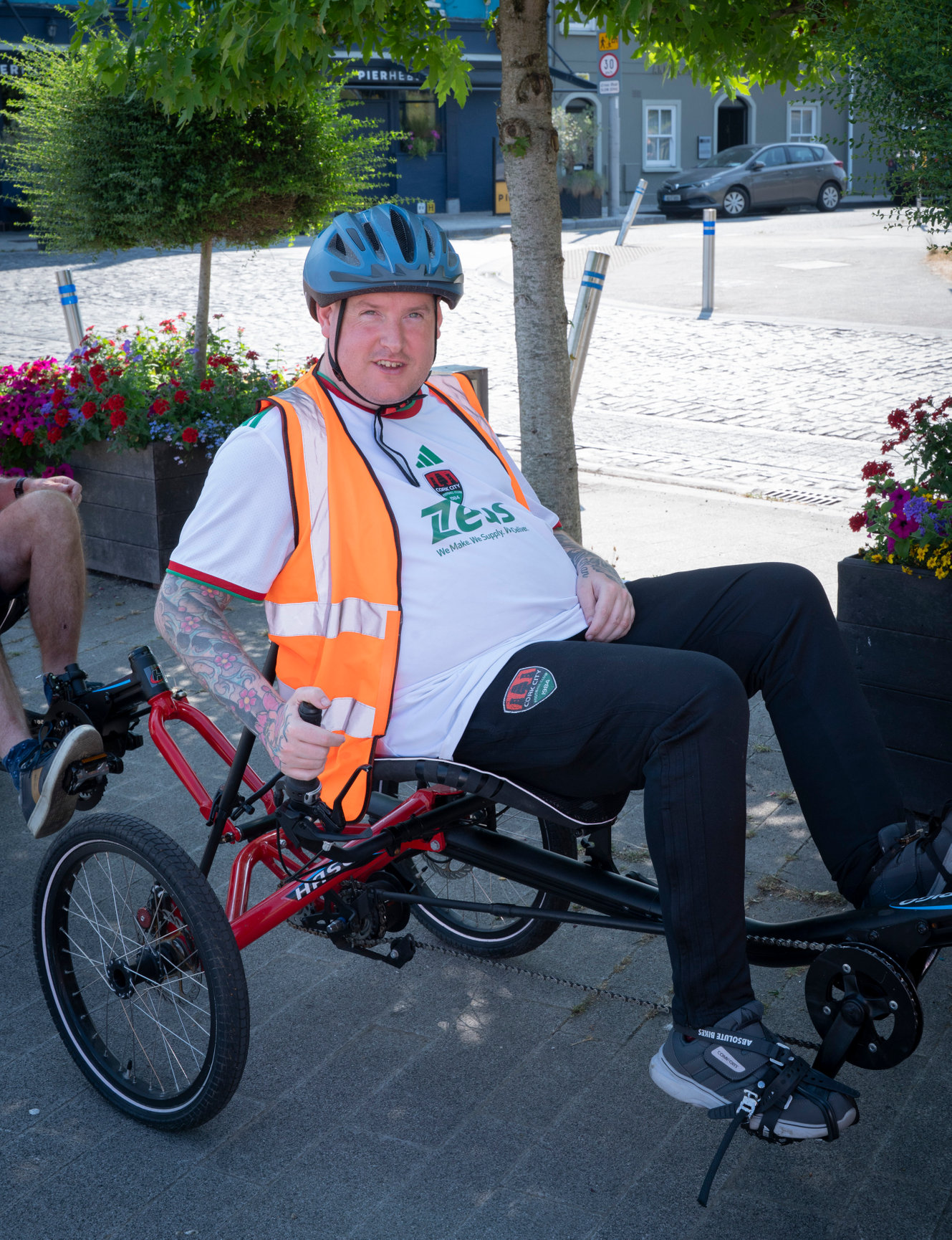 Life With No Limits Champion Jonathan taking part in an inclusive cycling event on an adapted recumbent bicycle, wearing a helmet and high‑visibility vest in a town centre setting.