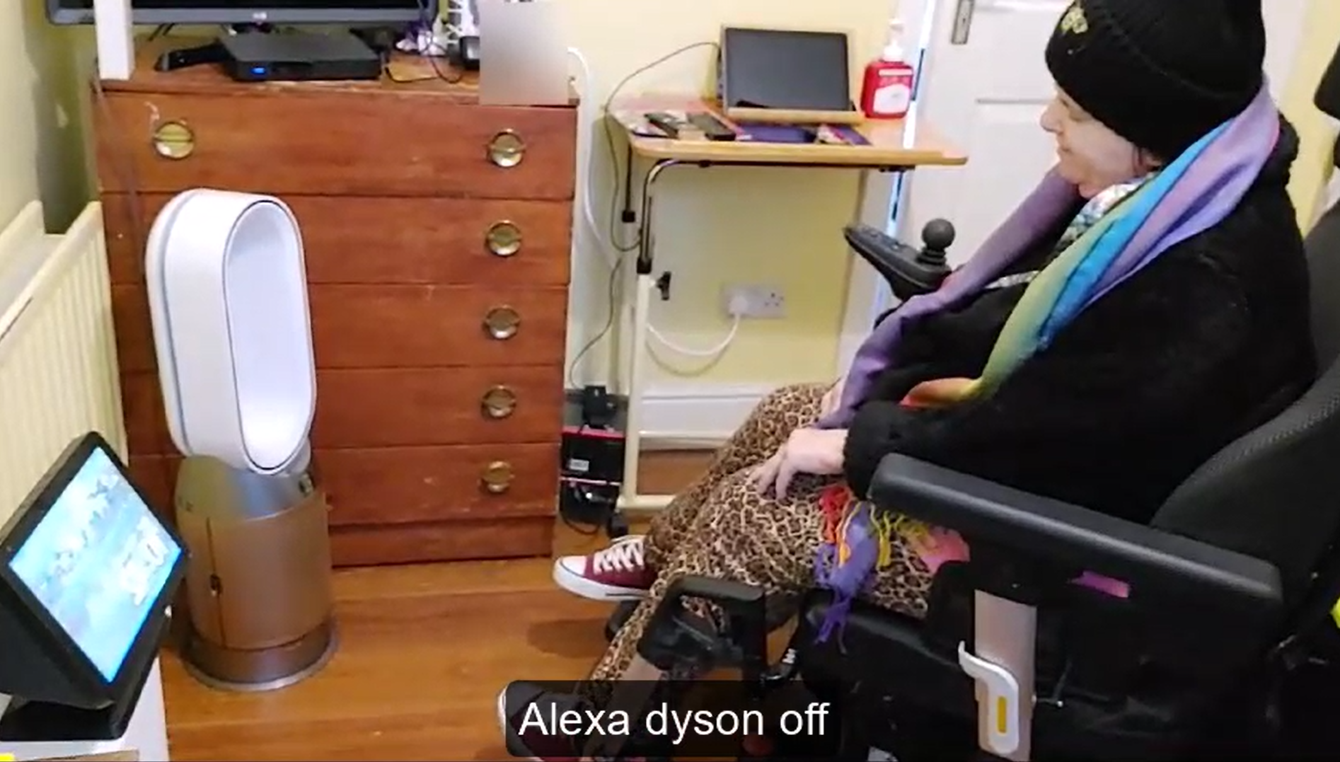 Person using a powered wheelchair in a smart home environment, controlling connected devices with voice technology. A smart display and smart fan are visible, demonstrating accessible technology that supports independent living.