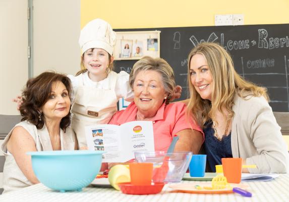 Three women and a child dressed as a chef gather around a table with colorful bowls and utensils, reading a pamphlet together.