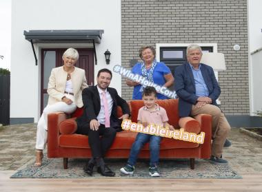 Four adults and a child sit on a couch in front of a house. They are holding signs which say #WinAHouseCork and #EnableIreland