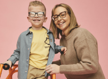Young child using an orange walking frame, standing beside an adult in a studio setting with a pink background, both facing forward in a supportive pose.