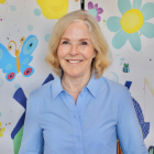 Enable Ireland Chair - Elizabeth J Barry A smiling woman with shoulder-length blonde hair, wearing a light blue button-up shirt and a pearl necklace. She is standing in front of a colorful, childlike mural featuring large flowers, a butterfly, and a bee.